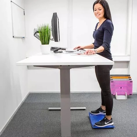 En kvinna står upprätt framför sitt skrivbord och jobbar, på en blå avlastningsmatta inne på kontoret.