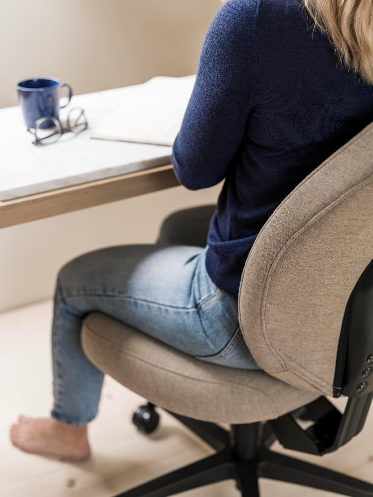 En kvinna klädd i jeans och en blå tröja sitter barfota vid sitt skrivbord på sitt hemmakontor, med en kaffekopp och glas på bordet.