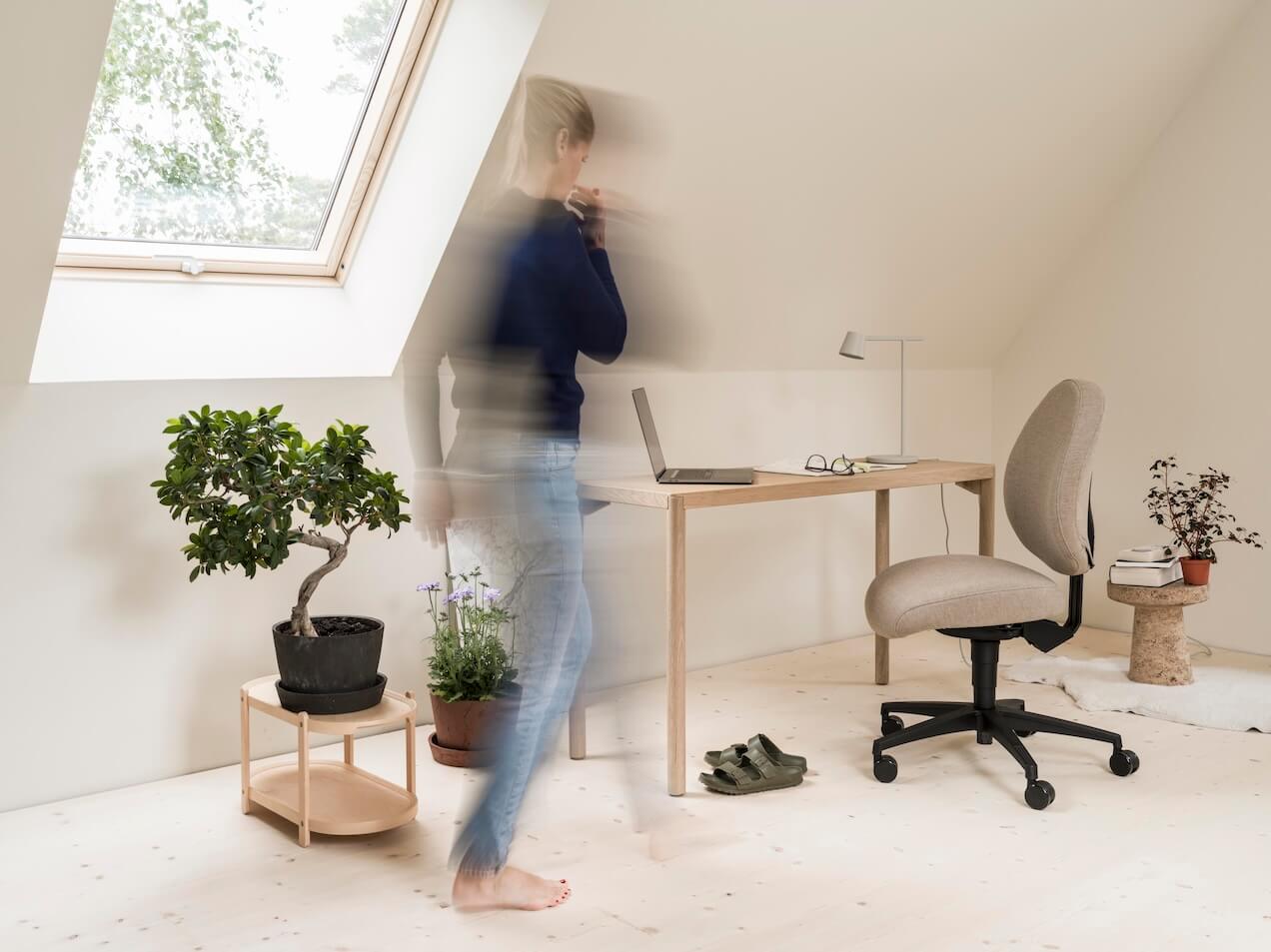 En kvinna är uppe och rör sig på sitt hemmakontor, med ett fönster, växter, ett bord med en arbetsstol i ett ljust vindsrum med sluttande tak.
